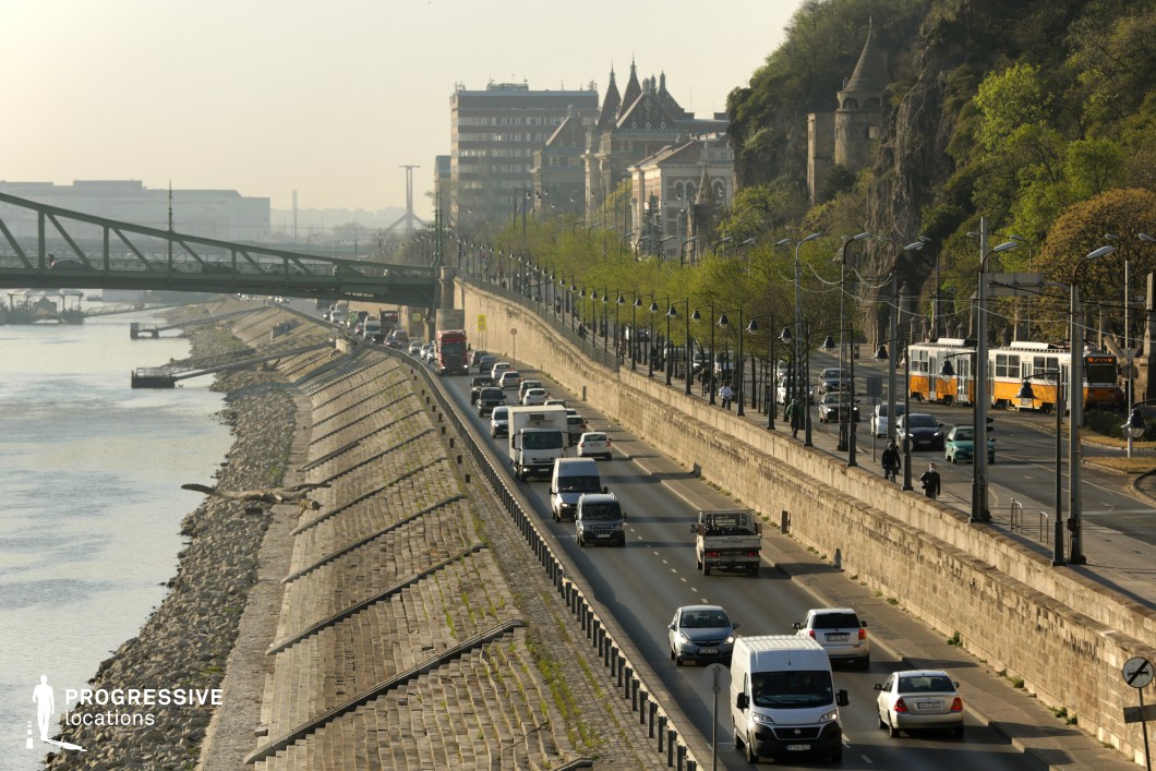 This image showcases a dynamic urban riverside scene where modern infrastructure meets historical elements. The multi-lane road runs parallel to the river, flanked by a steeply sloped stone embankment on one side and a row of vintage-style street lamps and lush trees on the other. A green metal bridge spans the water in the distance, connecting both sides of the city. On the right, a hillside with a historic stone turret-like structure emerges from the greenery, adding an old-world contrast to the contemporary traffic and tram system below. The composition's depth, with receding lines of vehicles, streetlights, and embankments, makes this an ideal location for film scenes requiring a visually rich European cityscape, suitable for chase sequences, travel montages, or atmospheric establishing shots.