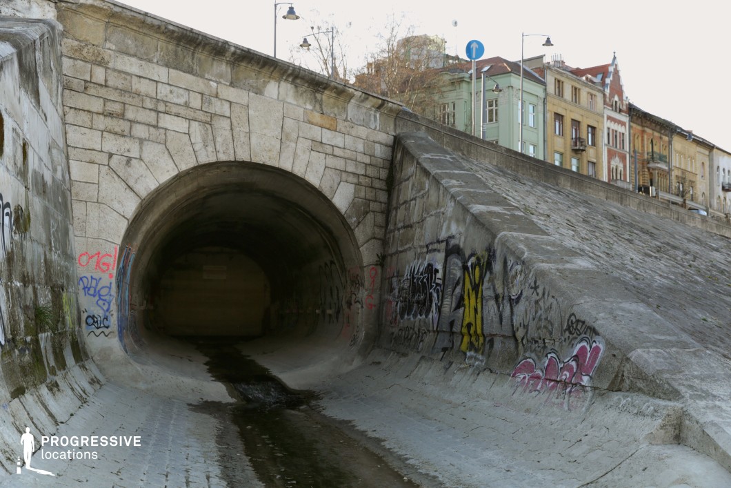 This image features a large, arched stone drainage tunnel embedded within a historic river embankment. The weathered stone blocks frame the tunnel entrance, which appears dark and mysterious, while graffiti adorns the surrounding concrete slopes. A trickle of water flows from the tunnel, carving a faint path along the textured ground. Above, a row of colorful, historic buildings with ornate facades lines the embankment, adding contrast to the scene. The location offers a gritty, urban aesthetic ideal for film settings requiring an underground passage, an urban escape route, or a secluded meeting spot.