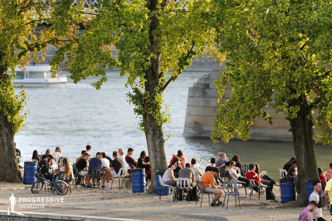 The image showcases a riverside café terrace, framed by leafy trees that provide shade and contribute to a relaxed atmosphere. Simple chairs line the path, arranged in a casual layout. The view of the river and the bridge in the distance adds a picturesque backdrop, enhancing the tranquil ambiance. This space, with its serene environment, is ideal for outdoor gatherings, leisure activities, or as a peaceful setting for urban scenes.