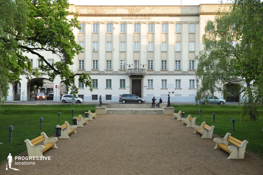 The image features a well-maintained park with a symmetrical arrangement of classic-design benches lining a gravel pathway leading to an imposing neoclassical-style building. The light-colored façade, adorned with decorative elements and a central balcony, suggests an institutional or governmental function. The park is framed by lush greenery, mature trees, and ornamental statues on pedestals, adding to the scene’s formal and elegant atmosphere. This location is ideal for filming historical dramas, political thrillers, or refined urban narratives, providing a structured and balanced setting for formal meetings, contemplative walks, or power dynamics between characters.