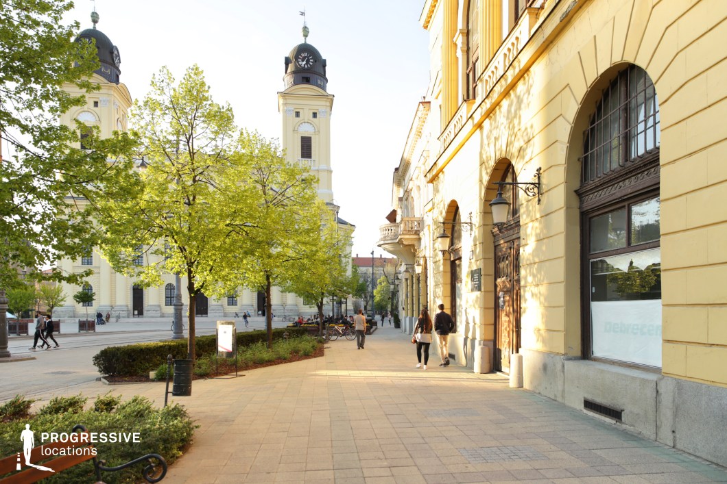 The image captures a charming European city square framed by elegant historic buildings and a grand clock tower with twin spires. The architecture is characterized by warm yellow facades, arched windows, and intricate detailing, evoking a classical and refined atmosphere. The paved walkway is lined with manicured greenery, ornate lampposts, and benches, adding to the inviting aesthetic. Sunlight filtering through the trees casts dynamic shadows, enriching the depth of the scene. This location is ideal for filming period dramas, romantic sequences, or travel-related productions, offering a visually rich and timeless setting for stories that emphasize history, elegance, or urban charm. The balanced composition allows for both wide establishing shots and intimate character moments.