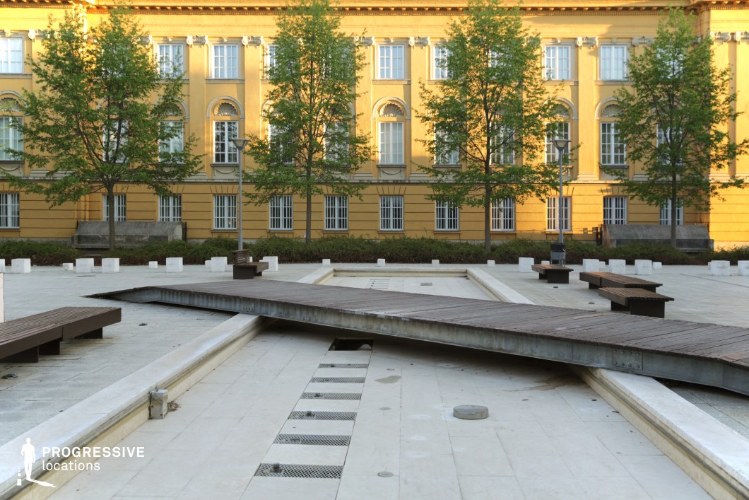 This image features a contemporary public square with a dry fountain structure, set against the backdrop of a stately yellow neoclassical building. The geometric arrangement of white stone elements and wooden pedestrian bridges creates a visually striking contrast with the historic architecture. The benches and trees add a structured rhythm to the space, enhancing its inviting yet minimalist atmosphere. The juxtaposition of modern and classical elements makes this location ideal for filming urban dramas, intellectual debates, or scenes that explore themes of history versus modernity, institutional authority, or academic life.