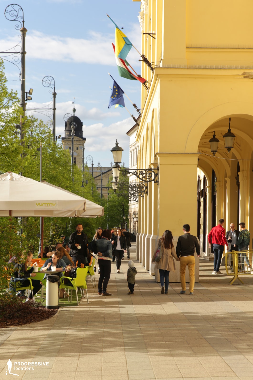 This image captures a lively city promenade lined with elegant yellow-arched buildings adorned with wrought-iron lanterns and flags. The paved walkway is complemented by outdoor café seating with bright modern furniture, contrasting with the classical architecture. In the background, a historic domed building and ornate street lamps add a European charm to the scene. The combination of architectural heritage and contemporary urban life makes this an excellent location for filming sophisticated city scenes, romantic encounters, or cultural narratives, offering a blend of history, elegance, and everyday vibrancy.