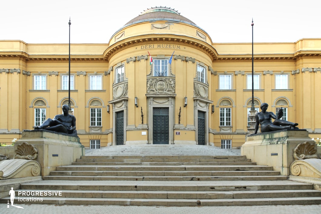 This imposing neoclassical museum façade features a symmetrical design with a central domed section, intricate sculptural reliefs, and large, ornate doors flanked by decorative lanterns. The warm yellow exterior is accentuated by arched windows and classical pilasters, reinforcing its historical elegance. A broad stone staircase leads up to the entrance, framed by two reclining bronze statues on pedestals, adding a monumental and timeless feel to the scene. This location is ideal for historical dramas, academic or mystery films, and political thrillers, offering a prestigious and authoritative setting for intellectual debates, institutional power struggles, or refined cultural encounters.