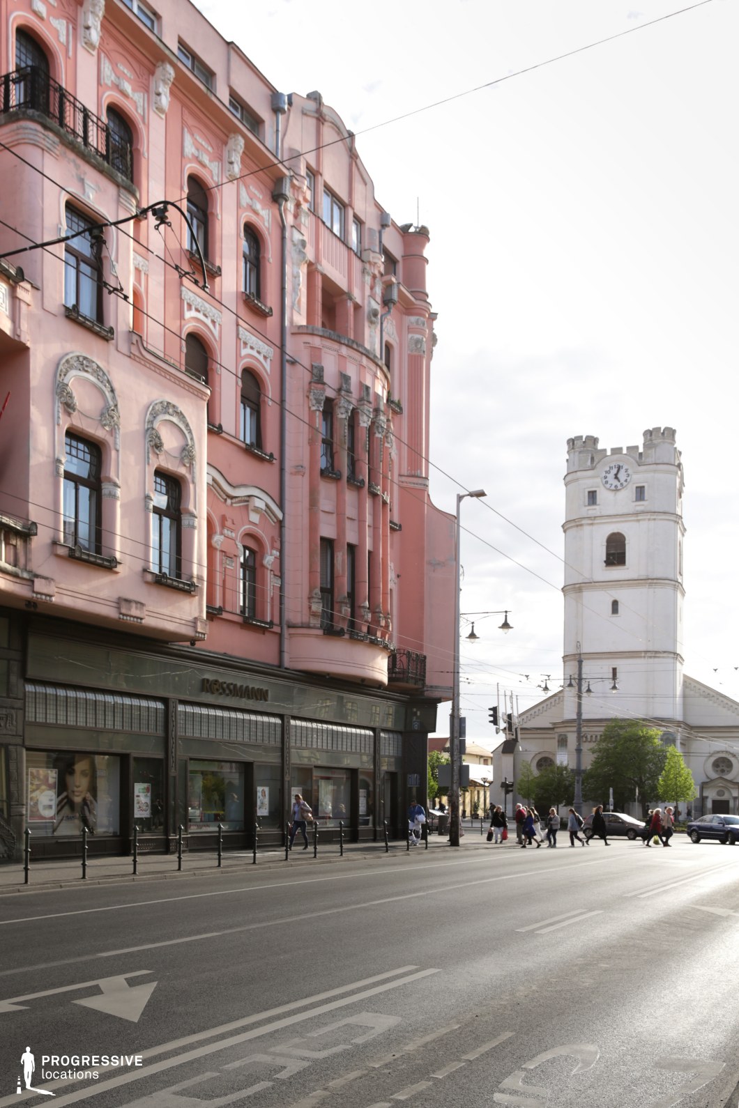This urban streetscape showcases a striking contrast between a richly decorated pink Secessionist-style building with intricate stucco details, arched windows, and wrought-iron balconies, and a stark white clock tower with a classical design in the background. The dynamic interplay of architectural styles, along with the tram cables cutting across the sky, creates a visually layered cityscape. The street-level storefronts and wide road enhance the scene’s metropolitan character. This location is ideal for filming period pieces, European city dramas, or urban stories requiring a visually compelling mix of historical elegance and contemporary street life.
