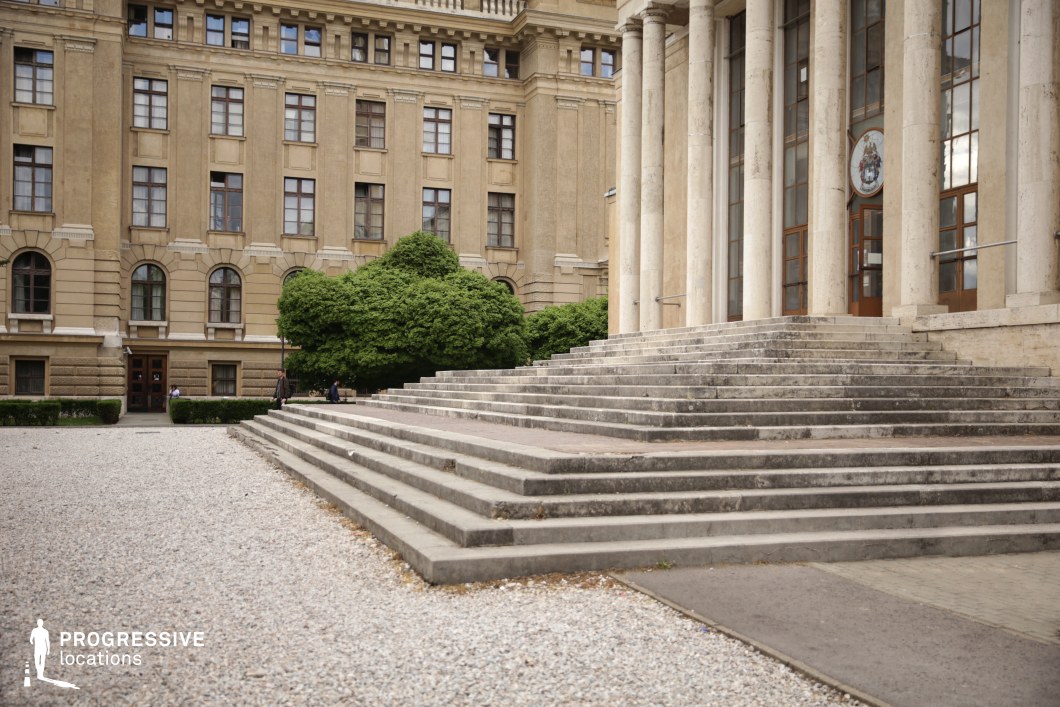 This imposing neoclassical-style building features a grand entrance marked by wide, weathered stone steps leading up to tall columns and large wooden doors with glass panels. The symmetrical façade, beige stone exterior, and detailed architectural elements suggest a historic government institution, university, or cultural center. The open courtyard, covered in gravel, enhances the sense of formality and grandeur, while the structured greenery softens the rigid geometry. This setting is ideal for filming academic dramas, political thrillers, or legal narratives, providing a dignified and authoritative backdrop for scenes involving power, tradition, or intellectual debate. The grand staircase also offers a visually compelling space for dynamic camera movements and dramatic confrontations.