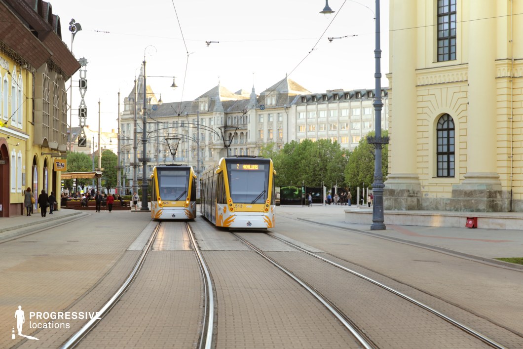 This city square blends historic and modern elements, featuring elegant neoclassical and late 19th-century buildings alongside contemporary yellow trams traveling along curved tracks. The cobblestone-paved streets, lined with traditional facades and decorative street lamps, evoke a European charm, while the open square is framed by lush green trees. The contrast between the ornate architecture and sleek tram design creates a visually engaging setting. This location is ideal for urban dramas, romantic narratives, or travel-themed productions, offering a dynamic backdrop for scenes set in a vibrant yet historically rich city center.