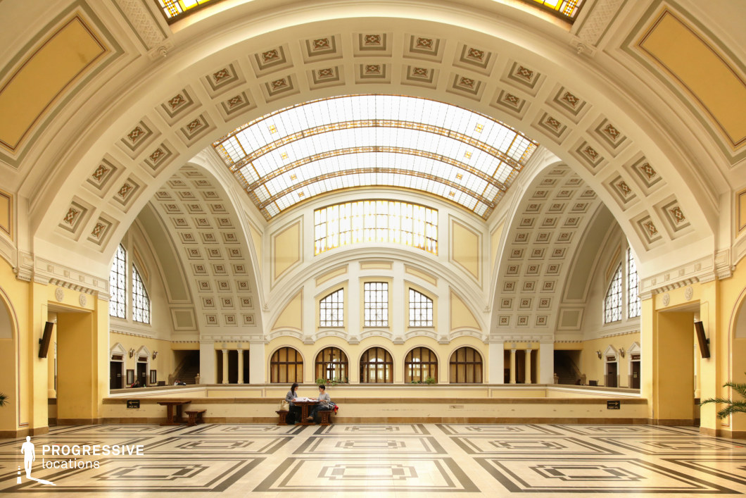 This image showcases a vast neoclassical-style hall with an impressive arched ceiling adorned with decorative geometric patterns. The high, curved windows and stained-glass skylights allow soft, diffused light to illuminate the space, enhancing the refined elegance of the pale yellow and white interior. The floor features a symmetrical black-and-white geometric tile pattern, adding to the sense of grandeur. The hall’s arched openings, classical detailing, and expansive proportions make it an ideal location for filming historical dramas, period pieces, academic or institutional settings, and refined, high-society narratives. Its stately atmosphere also lends itself to themes of mystery, authority, or intellectual discourse.