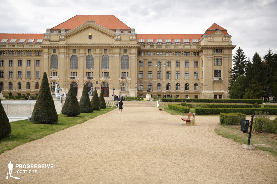 This stately neoclassical building, framed by a meticulously landscaped garden, features an elegant symmetrical facade with grand arched windows, decorative columns, and a central pediment. The wide gravel path leading to the entrance is lined with manicured hedges, sculpted trees, and classical statues, enhancing the refined, academic atmosphere. The combination of historic architecture and formal garden design creates a prestigious and cinematic setting. This location is ideal for filming period dramas, intellectual thrillers, or coming-of-age stories, serving as an academic institution, government building, or aristocratic estate backdrop for scenes of scholarly discussions, political intrigue, or contemplative walks.