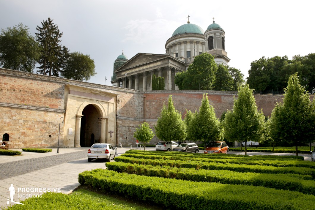 This grand basilica, with its neoclassical domes and towering columns, stands majestically above fortified brick walls, creating a visually striking blend of religious and defensive architecture. The lower section features a historic arched gateway, leading into a well-manicured garden with neatly trimmed hedges and symmetrically planted trees. The contrast between the aged, textured brickwork and the refined stone facade enhances the location’s depth and historical significance. This setting is ideal for period dramas, historical epics, and architectural documentaries, providing an atmospheric backdrop for regal, spiritual, or medieval-themed productions.
