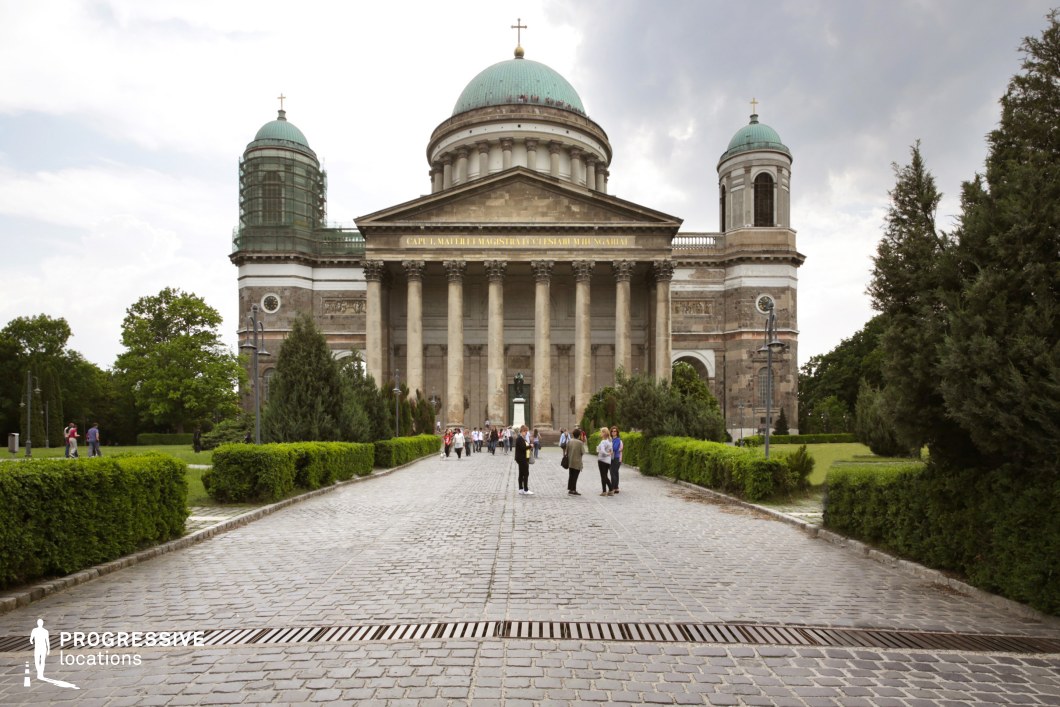 This image showcases a monumental neoclassical basilica with towering Corinthian columns, an expansive dome, and two symmetrical bell towers. The aged stone facade, featuring intricate details and Latin inscriptions, exudes historical and architectural significance. The basilica is approached via a long, straight cobblestone promenade lined with manicured hedges and lampposts, reinforcing the site's grandeur and formality. The surrounding greenery contrasts with the imposing structure, adding depth to the setting. This location is ideal for filming historical dramas, religious narratives, or epic period pieces, offering a stately and timeless European atmosphere.