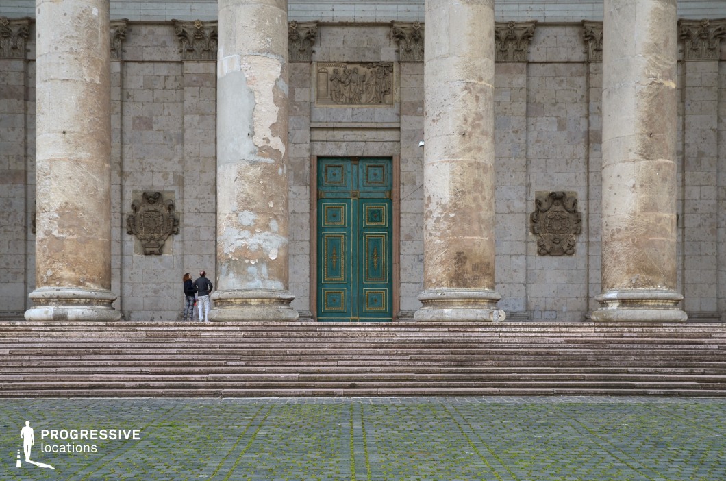 This imposing neoclassical entrance features towering, weathered stone columns that frame a large, ornate green door with intricate gold detailing. The aged stone façade, adorned with decorative reliefs and coats of arms, adds a sense of historical depth and grandeur. The broad, worn steps leading to the entrance reinforce the building’s monumental scale, while the moss-lined cobblestone foreground introduces a touch of organic contrast to the rigid architecture. This location is ideal for filming historical dramas, religious or political thrillers, and narratives involving power, secrecy, or legacy. The grand scale and timeless aesthetic provide a cinematic backdrop for scenes of authority, revelation, or confrontation.