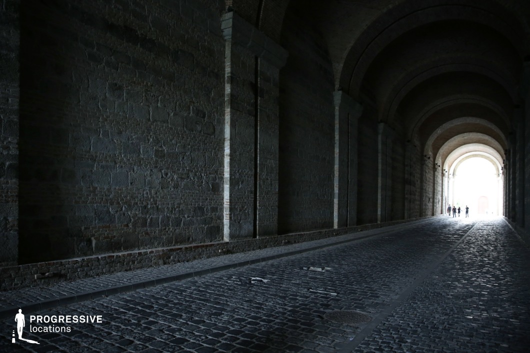 The image features a long, atmospheric tunnel with massive stone walls and a series of grand arches that create a sense of depth and historical gravitas. The dark cobblestone road, worn and uneven, enhances the aged character of the passage. The interplay of light and shadow emphasizes the textured surfaces, with the tunnel’s exit appearing as a bright, almost ethereal contrast at the far end. This location is ideal for filming historical epics, suspenseful period dramas, or noir-style thrillers, offering a striking setting for mysterious pursuits, secret rendezvous, or dramatic entrances and exits.