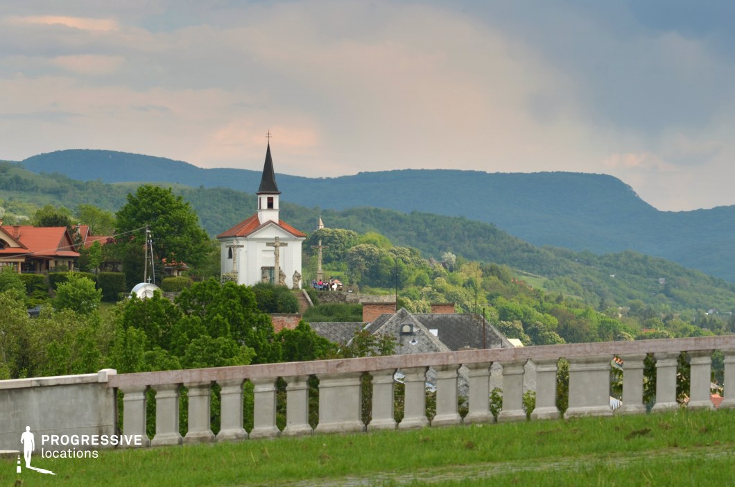 The image captures a picturesque hillside village with a small white church featuring a red roof and a pointed steeple, nestled among lush greenery. The surrounding landscape consists of rolling hills covered in dense forests, creating a layered natural backdrop. Traditional houses with pitched roofs and rustic elements add to the rural charm of the scene. A stone balustrade in the foreground frames the composition, adding a touch of classical elegance to the view. This location is ideal for filming historical dramas, rural life stories, or contemplative and romantic scenes. The serene setting and harmonious blend of architecture and nature make it well-suited for productions emphasizing tradition, nostalgia, or personal introspection.