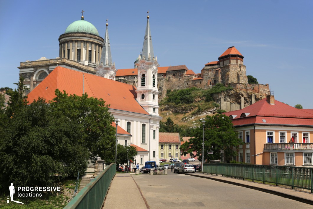 This stunning European cityscape features a majestic basilica with a grand dome, an elegant twin-spired church, and a historic hilltop castle overlooking the scene. The combination of Baroque and Renaissance architectural elements, red-tiled rooftops, and pastel-colored buildings creates a visually rich and timeless setting. The bridge in the foreground leads toward the heart of the city, enhancing depth and perspective. This location is ideal for filming historical dramas, period epics, or romantic narratives, offering an authentic old-world ambiance that evokes cultural heritage, grandeur, and historical significance.