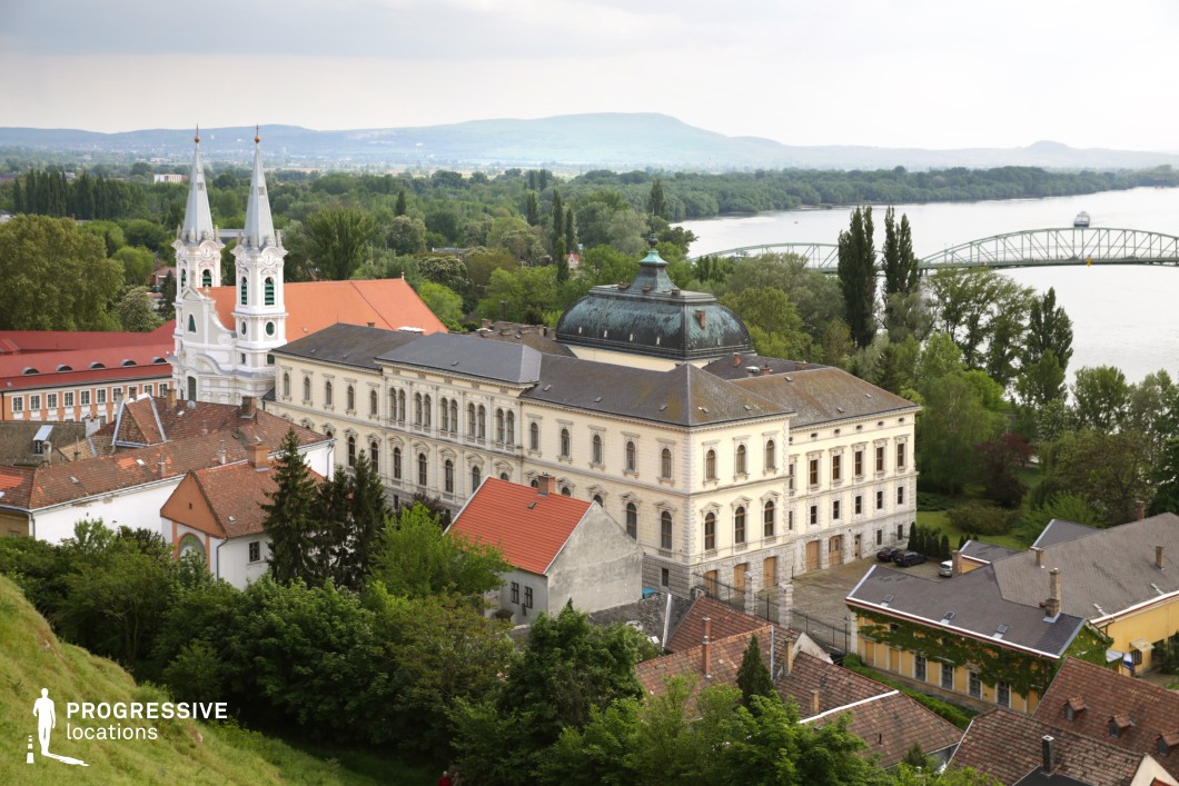 This panoramic view showcases a picturesque European town nestled along a winding river, featuring an elegant neoclassical building with a domed roof and a striking white church with twin spires. The rooftops of charming historic houses, covered in red tiles, contrast beautifully against the lush greenery and rolling hills in the background. A gracefully arched bridge spans the river, adding to the scenic composition. The timeless quality of the architecture and the tranquil waterside setting make this location ideal for filming historical dramas, period romances, or cultural documentaries, offering a visually rich backdrop for narratives of heritage, diplomacy, or poetic nostalgia.