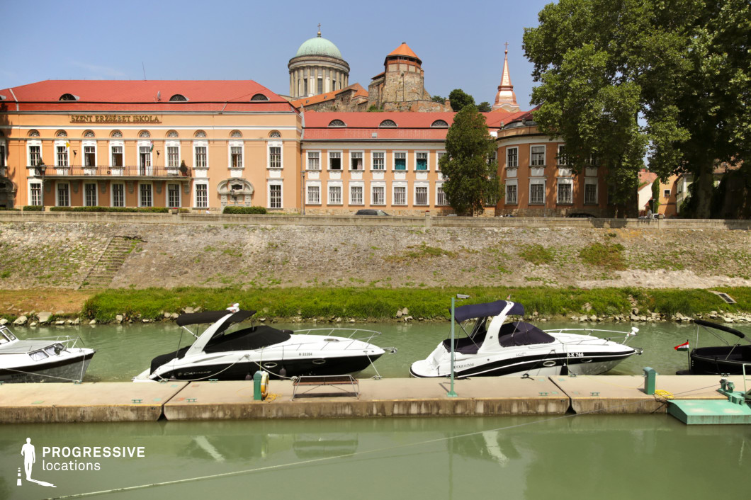 This picturesque riverside setting features a small marina with sleek motorboats moored along a calm waterway, contrasting with the historic architecture in the background. The buildings showcase a blend of classical and baroque influences, with warm-toned facades, arched windows, and decorative details. A prominent domed structure and a fortified tower add grandeur, hinting at the location's historical significance. The sloped stone embankment creates a natural separation between the water and the cityscape, enhancing the scene’s visual depth. This location is ideal for filming elegant period dramas, travel documentaries, or narratives centered around affluent lifestyles, political intrigue, or historical discoveries.