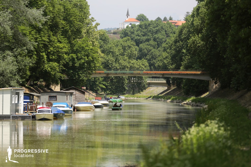 This image captures a peaceful riverside scene lined with small boats moored along rustic wooden docks and floating structures. The river is bordered by lush green trees that frame the winding waterway, leading towards a distant bridge and a picturesque hilltop church with a red roof. The mix of natural elements and man-made structures, including a second, ornate bridge in the background, adds depth and character to the setting. This location is ideal for filming reflective, scenic narratives, rural dramas, or slow-paced adventure stories, offering a timeless, tranquil backdrop for contemplative moments or quiet riverside life.