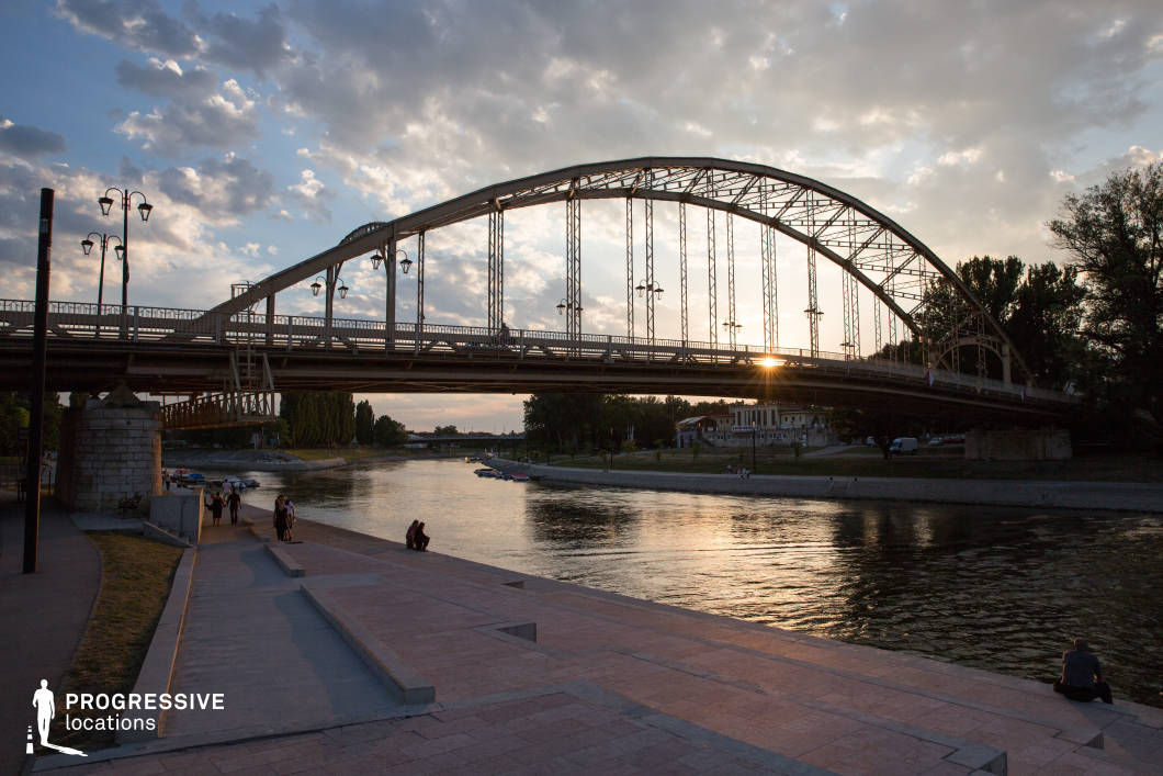 This image features a graceful arched steel bridge spanning across a calm river, with a well-designed riverside promenade in the foreground. The structured pedestrian walkway includes tiered steps leading to the water’s edge, providing a dynamic visual depth. The bridge’s intricate metal framework, lined with evenly spaced lampposts, adds a classic and elegant industrial touch. The riverbank is dotted with small boats and historic buildings, creating a harmonious urban atmosphere. This location is ideal for filming romantic dramas, introspective narratives, or urban adventure films, offering a scenic and cinematic setting with strong compositional symmetry.