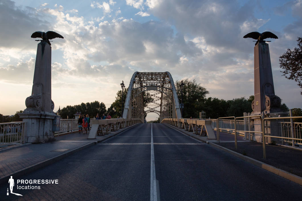 This striking steel arch bridge features an elegant lattice framework, flanked by two imposing stone pillars adorned with sculpted eagles. The symmetrical design and central roadway create a sense of depth and perspective, drawing the eye toward the horizon. Ornate street lamps line the pedestrian walkway, enhancing the bridge’s historic character. The combination of steel and stone elements gives the structure a strong, enduring presence. This location is ideal for dramatic, action-packed, or introspective scenes in road movies, historical dramas, or thrillers, offering a visually compelling setting for moments of transition, confrontation, or escape.