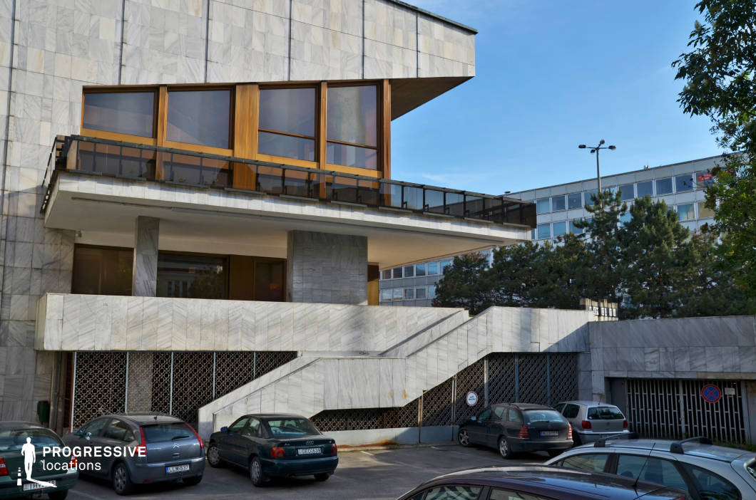 The image features a striking brutalist-style building with a distinct geometric design, clad in large marble panels that give it a refined yet imposing presence. The structure incorporates sharp angular lines, an elevated terrace with a glass railing, and large wooden-framed windows that contrast with the cool stone facade. A wide, sculptural staircase leads to the upper level, while a patterned metal lattice encloses the lower section, adding depth and texture. The surrounding environment includes mid-century modern office buildings and greenery, enhancing the urban aesthetic. This location is ideal for filming political dramas, corporate thrillers, or neo-noir narratives, offering a visually striking setting that conveys power, authority, or institutional rigidity. Its architectural boldness and minimalist elegance make it suitable for scenes requiring a modernist or dystopian backdrop.