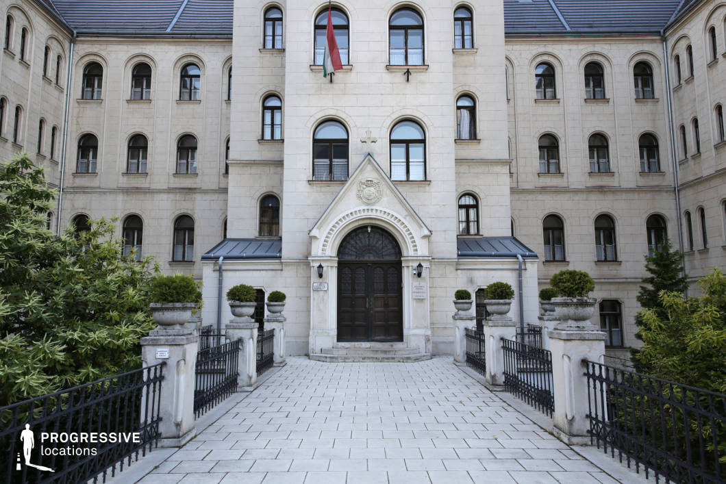 The image features an imposing neoclassical-style building with a symmetrical façade, arched windows, and a grand central entrance framed by an ornate triangular pediment. A stone-paved pathway leads to the main doors, flanked by decorative planters and wrought-iron railings, enhancing the stately and formal atmosphere. The light-colored stone exterior contrasts with the darker roof and wooden doors, emphasizing the structure’s historic character. This location is ideal for filming period dramas, academic or governmental settings, or mystery films, serving as an authoritative backdrop for university scenes, legal institutions, or secretive meetings in a historical context.