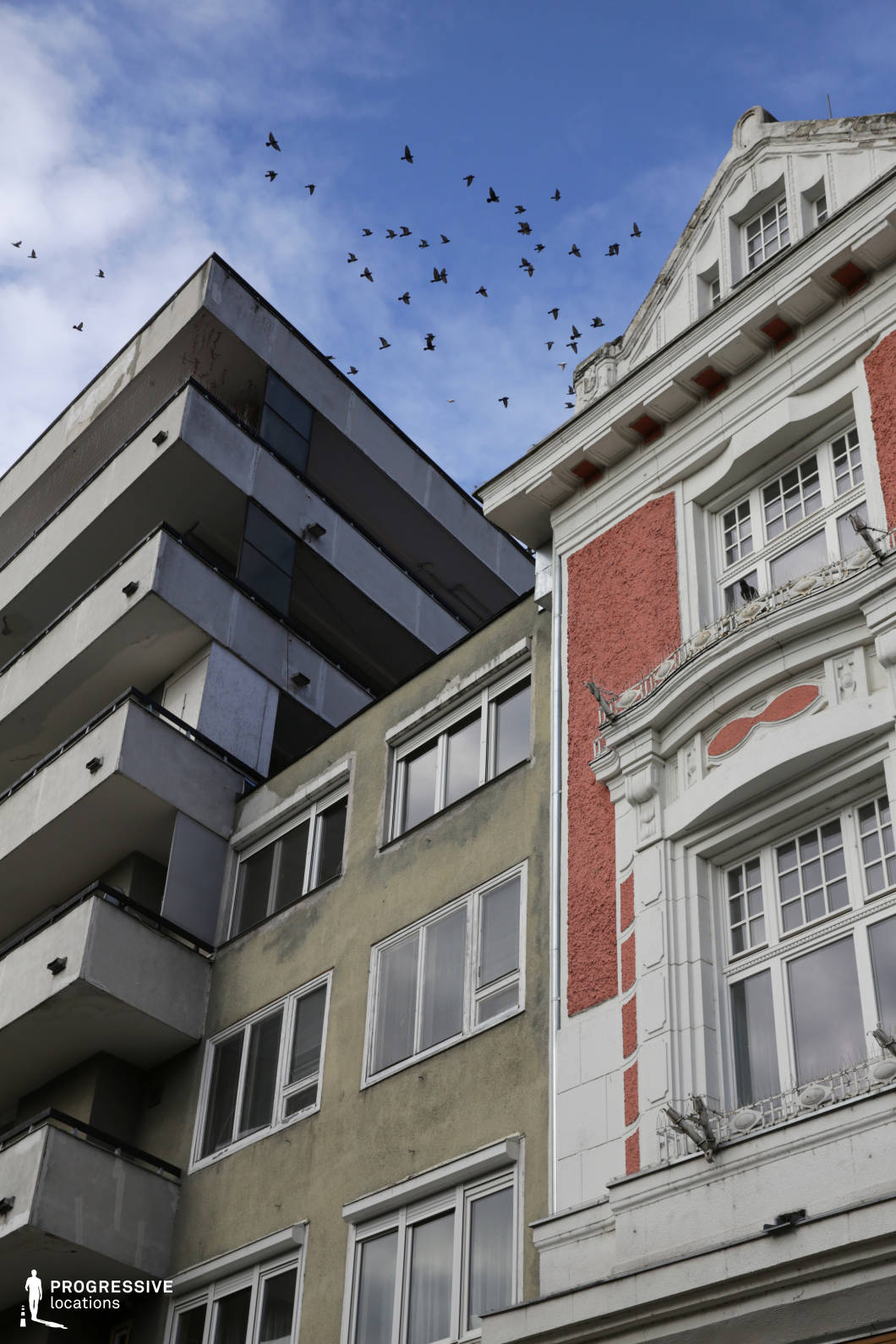This image captures a striking juxtaposition of architectural styles, with a highly ornamented historic building standing adjacent to a stark, modernist concrete apartment block. The ornate facade features intricate detailing, curved windows, and decorative moldings, while the adjacent structures exhibit weathered surfaces and geometric rigidity, emphasizing urban decay. A flock of birds soaring above adds a dynamic element, reinforcing the passage of time and the contrast between old-world elegance and brutalist functionality. This location is ideal for filming urban dramas, psychological thrillers, or social commentaries, offering a visually compelling backdrop that conveys themes of change, nostalgia, and societal contrast.