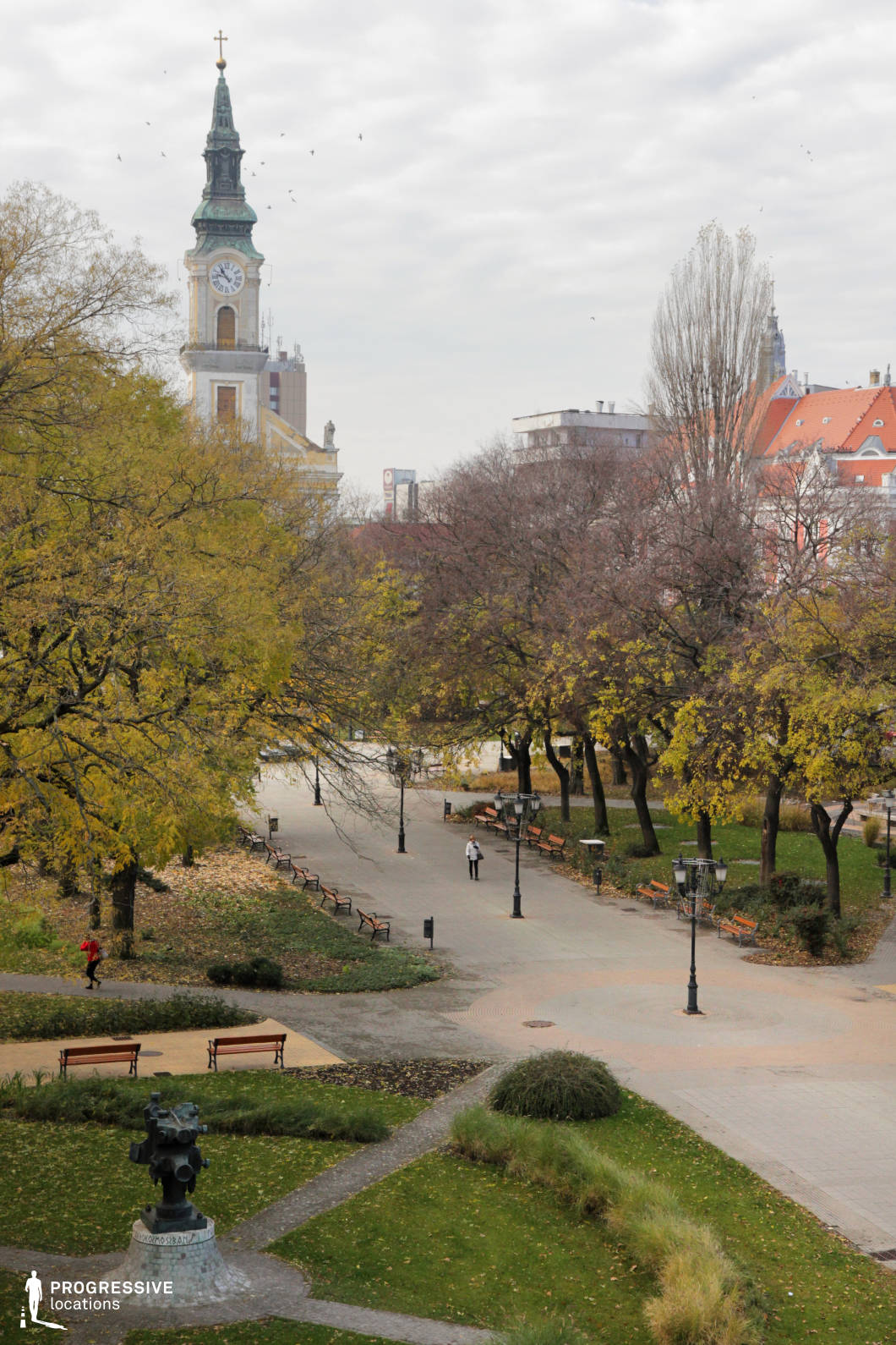 This image captures a picturesque European town square featuring a landscaped park with winding pathways, manicured greenery, and classical lampposts. The centerpiece is an elegant historic church with a tall, ornate clock tower, its Baroque architectural details standing in contrast to modern buildings in the background. The composition balances nature and architecture, with tree-lined walkways and scattered benches enhancing the inviting atmosphere. The location is ideal for filming historical dramas, romantic scenes, or contemplative narratives, offering a visually rich setting that blends cultural heritage with an urban park environment.