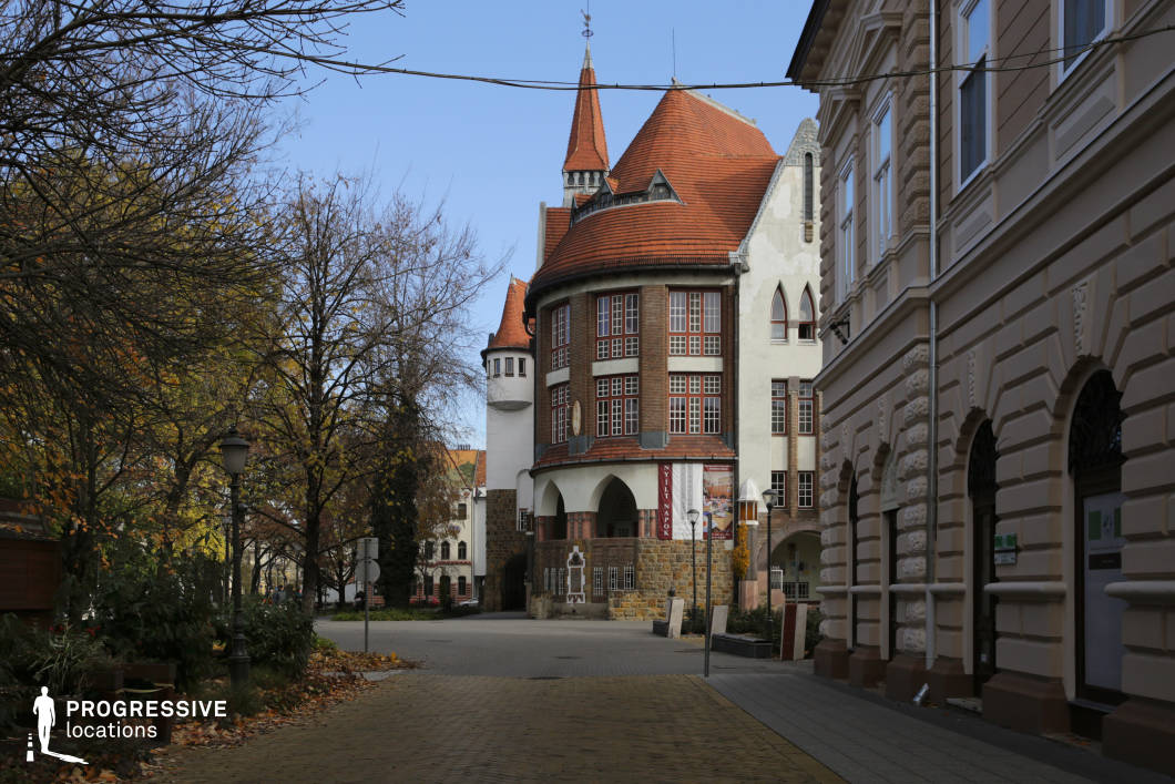 This distinctive architectural landmark features a blend of medieval and Art Nouveau elements, with a rounded tower, pointed spires, and decorative half-timbered windows. The steep red-tiled roofs and arched entranceways enhance the historic character, giving the structure a castle-like presence within an urban setting. The surrounding cobbled streets, classic street lamps, and adjacent buildings with detailed façades create a charming and immersive atmosphere. This location is ideal for filming period dramas, fantasy stories, or historical mysteries, offering a visually striking and whimsical backdrop. The mix of architectural styles and warm tones makes it suitable for narratives involving intrigue, nostalgia, or cultural heritage.