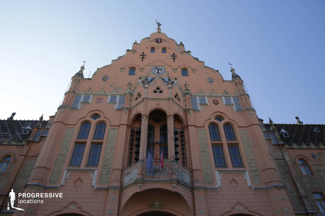 This grand Art Nouveau-style town hall features an elaborate, asymmetrical facade with intricate detailing, colorful heraldic motifs, and arched windows that emphasize its decorative character. The pastel-toned exterior, adorned with sculpted elements, ceramic tiles, and a central balcony, creates a striking visual presence. A prominent clock and ornamental gables add to the historic grandeur of the structure. The upward perspective enhances the building’s imposing stature, making this location ideal for filming period dramas, political thrillers, or historical documentaries that require a majestic and architecturally rich setting.