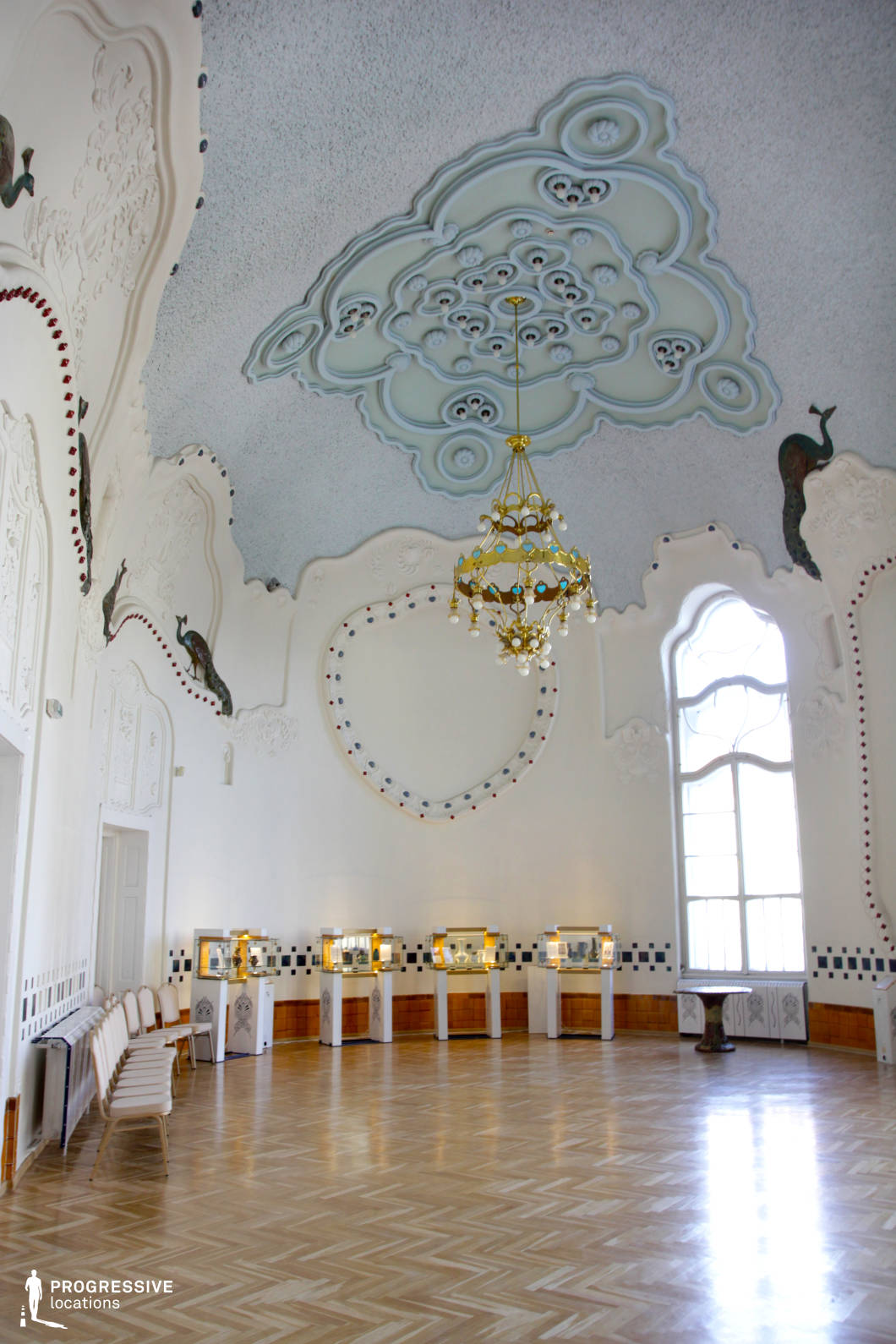 This exquisite Art Nouveau hall features an intricately designed ceiling with organic, flowing patterns in soft pastel hues, accompanied by sculptural peacock motifs along the walls. The large arched window allows natural light to highlight the elegant parquet flooring and delicate decorative elements. The golden chandelier adds a touch of sophistication, reinforcing the opulent yet whimsical atmosphere. This location is ideal for filming period dramas, artistic productions, or fantasy narratives, offering a dreamlike setting for refined social gatherings, historical storytelling, or visually captivating scenes centered on elegance and cultural heritage.