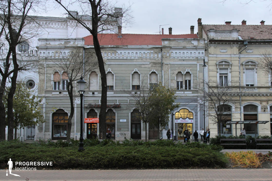 The image showcases a row of elegant historic buildings with intricate architectural details, situated along a tree-lined street. The facades feature a mix of arched windows, decorative cornices, and ornamental plasterwork, though some areas show signs of aging and wear. The ground level houses various small shops with inviting storefronts, while a bus stop with a modern glass shelter integrates contemporary elements into the scene. In the foreground, a well-maintained park with benches, lampposts, and greenery adds depth and contrast to the structured urban environment. This location is ideal for filming period dramas, cultural documentaries, or urban storytelling, offering a refined yet slightly weathered European ambiance suited for historical narratives, character-driven dramas, or leisurely city explorations.