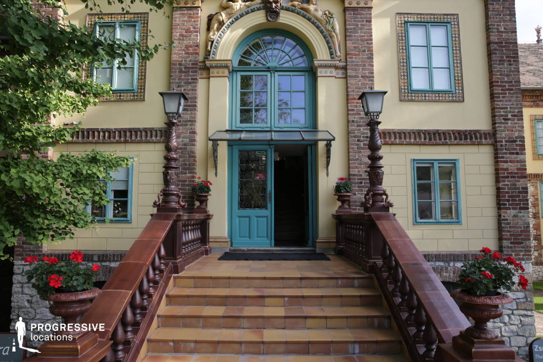 The image showcases an elegant entrance to a historic building, featuring a grand tiled staircase flanked by dark wooden balustrades and intricate lampposts. The façade blends brickwork and pastel-colored plaster, with decorative window frames and ornamental reliefs adding character. The arched doorway, painted in a striking turquoise hue, is adorned with sculptural elements that enhance its Art Nouveau charm. This location is ideal for filming period dramas, upscale historical narratives, or whimsical fantasy settings, providing a richly detailed and visually captivating backdrop for grand entrances, aristocratic settings, or mysterious encounters.