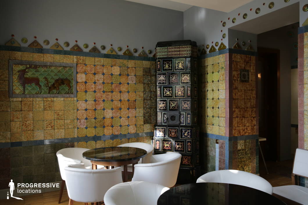 This richly decorated interior showcases a striking Art Nouveau aesthetic, with intricate ceramic tilework covering the walls in warm earth tones, geometric patterns, and nature-inspired motifs. A prominent black ceramic stove with ornate embellishments serves as a focal point, contrasting with the surrounding decorative surfaces. The room is furnished with modern white chairs and sleek black tables, blending contemporary comfort with historic craftsmanship. This location is ideal for filming scenes that require an artistic, vintage, or culturally refined atmosphere, making it suitable for period dramas, sophisticated dialogues, or intimate café settings with a distinctive visual identity.