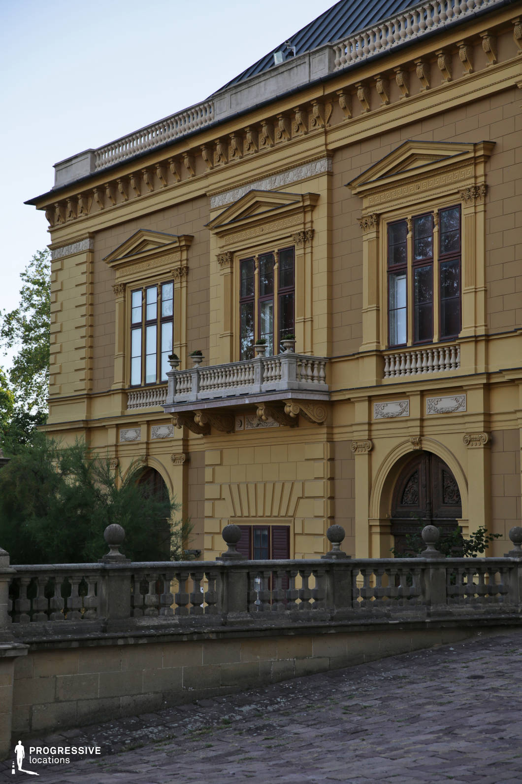 This grand neoclassical mansion features a richly detailed façade with symmetrical window arrangements, decorative pediments, and intricate stonework. A prominent sculpted balcony with balustrades adds a regal touch, complemented by a stately arched entrance below. The ochre-colored exterior enhances the building’s historic and aristocratic character. In the foreground, a stone balustrade and a gently sloping cobblestone path contribute to the scene’s old-world sophistication. This location is ideal for period dramas, aristocratic narratives, or historical intrigue, providing a refined and visually compelling backdrop for scenes of power, romance, or intrigue.