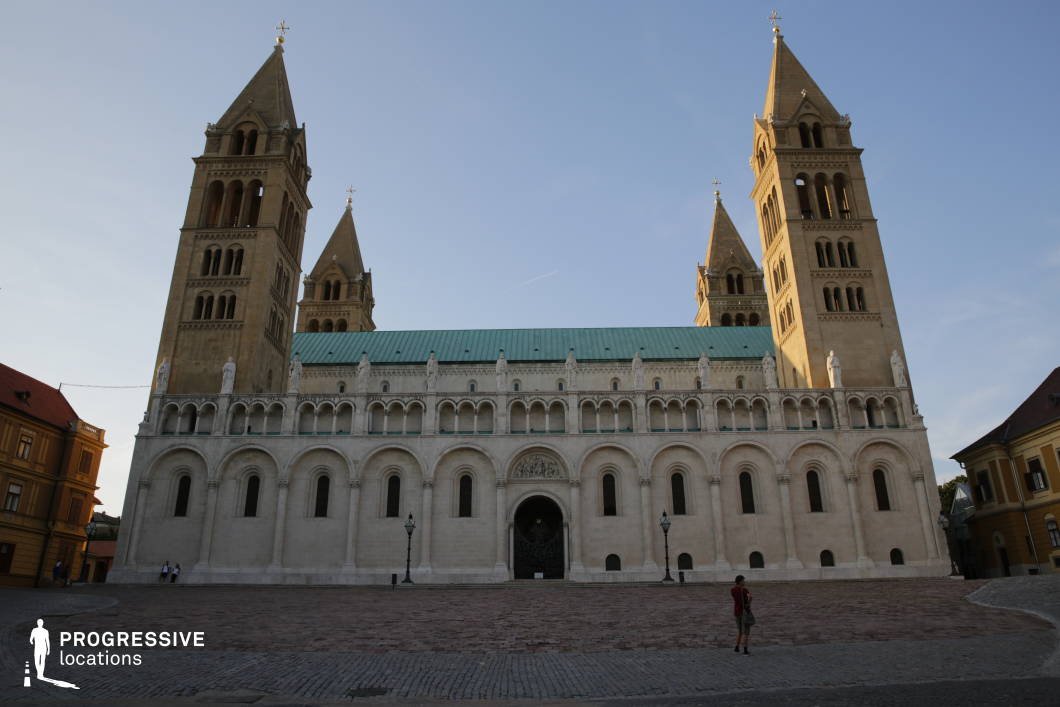 This monumental Romanesque cathedral stands with imposing symmetry, featuring four towering spires, a series of rounded arches, and a strikingly clean stone facade. The green copper roof contrasts with the warm tones of the surrounding historic buildings, while the cobblestone plaza in the foreground enhances the grandeur of the structure. Ornate statues and intricate carvings adorn the upper sections, adding depth to its historical and architectural significance. This location is ideal for filming period dramas, historical documentaries, or epic narratives, offering a majestic and timeless setting for scenes depicting power, faith, or medieval heritage.