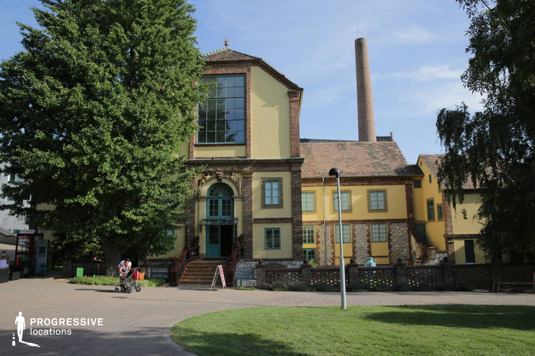 This image showcases a distinctive historic industrial building with a mix of brick, stone, and plaster elements, blending functional architecture with decorative detailing. The structure features large arched windows, intricate brickwork, and a central entrance framed by an elaborate doorway with classical embellishments. The towering chimney in the background reinforces its industrial heritage, while the surrounding landscaped park with paved walkways and mature trees softens the setting. This location is ideal for filming historical dramas, industrial-era period pieces, or creative reinterpretations of repurposed spaces, offering a unique mix of past craftsmanship and modern adaptation.