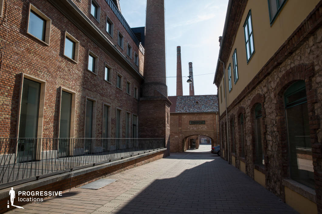 This narrow industrial alley is framed by historic brick and stone buildings, leading to an arched gateway that opens into a larger factory complex. Towering smokestacks dominate the skyline, hinting at the site's former industrial purpose. The combination of red brick facades, modernized windows, and iron railings creates a blend of past and present, reinforcing the site’s adaptive reuse. The long shadows and enclosed passageway add depth and intrigue. This location is ideal for filming historical dramas, noir-inspired thrillers, or industrial-era narratives, offering an atmospheric setting for clandestine meetings, urban chases, or explorations of the past.