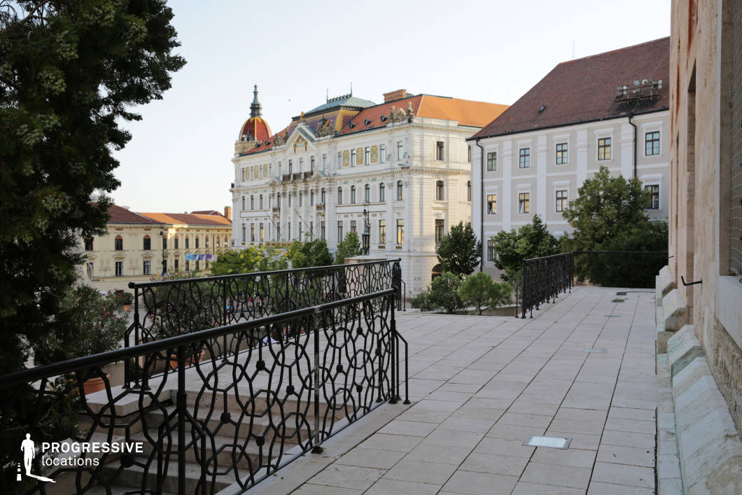 This elevated terrace overlooks a grand historic square, offering a stunning view of ornate neoclassical and baroque buildings adorned with intricate detailing, domed rooftops, and decorative balconies. The wrought-iron railing and stone-paved terrace create a sophisticated foreground, adding depth to the composition. The surrounding greenery enhances the contrast between architectural grandeur and natural elements. This location is ideal for filming period dramas, political thrillers, or romantic narratives, providing a refined and visually dynamic backdrop. The elevated perspective makes it well-suited for contemplative moments, high-stakes conversations, or scenes depicting power and prestige.