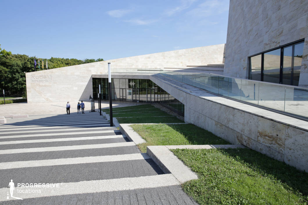This sleek, modern concert hall showcases a minimalist architectural design with angular, geometric shapes and a facade clad in light-colored stone. The expansive glass windows and transparent railings create a seamless connection between the structure and its surrounding greenery. The wide, striped stairway and clean lines add to the building’s refined and sophisticated aesthetic. This location is ideal for filming cultural documentaries, high-end corporate scenes, or artistic dramas, providing a striking and contemporary backdrop for moments of creative expression, intellectual discourse, or elegant public events.