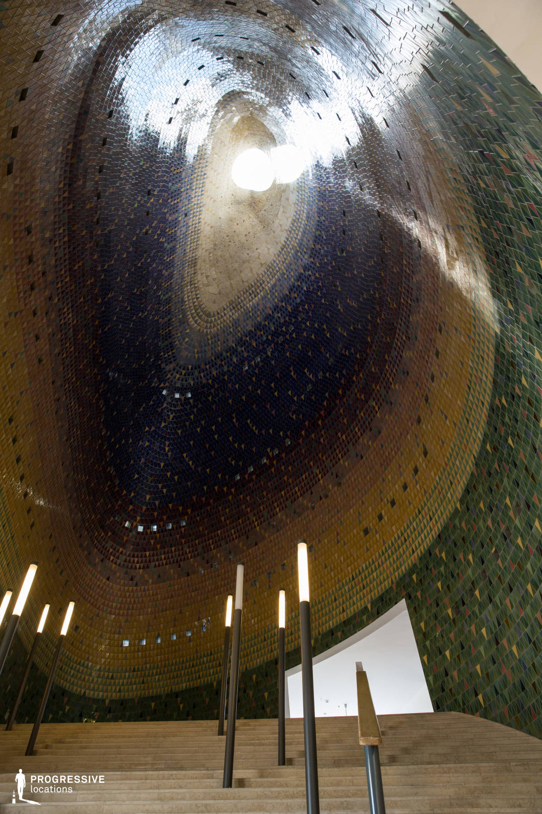 This striking architectural space features a sweeping mosaic dome adorned with an intricate gradient of green, gold, and deep blue tiles, culminating in a sculptural skylight that allows natural light to pour in. The grand staircase leading upward is flanked by minimalist, glowing light fixtures, creating a futuristic yet organic ambiance. The curvature of the dome, combined with the rich texture of the tiles, gives the space a cavernous yet intimate feel, resembling a modern sanctuary or cultural venue. This location is ideal for sci-fi, fantasy, or art-house productions, serving as a backdrop for visionary sequences, intellectual debates, or moments of transcendence in an avant-garde setting.