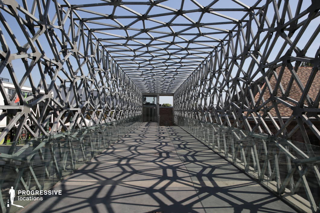 This striking pedestrian bridge features an intricate lattice-like steel framework that creates a dynamic interplay of light and shadow on the walkway below. The geometric patterns extend across the entire structure, giving it a futuristic, almost sculptural quality. The transparent glass railings enhance the sense of openness while providing an unobstructed view of the surroundings. The combination of industrial materials and innovative design makes this location ideal for filming modern thrillers, sci-fi narratives, or high-stakes action sequences. The bridge’s repeating patterns and vanishing point perspective also lend themselves to visually engaging cinematography, making it a compelling setting for dramatic or symbolic moments.