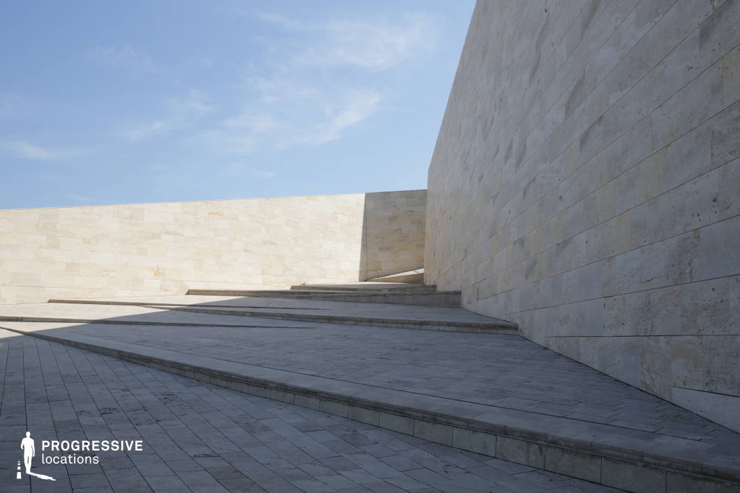 The image features a striking minimalist architectural composition dominated by large, pale stone walls and sloping walkways. The geometric design creates a sense of scale and depth, with clean lines leading the eye toward the vanishing point. The smooth, uniform stone surfaces contrast with the sharp angles of the steps and ramps, enhancing the modernist aesthetic. Shadows cast by the walls add dimension, emphasizing the stark, almost surreal quality of the space. This location is ideal for filming contemplative, futuristic, or abstract sequences, making it well-suited for sci-fi, dystopian, or high-concept artistic productions. The vast openness and absence of ornamentation create a neutral yet dramatic setting for scenes involving solitude, decision-making, or transitions between spaces.