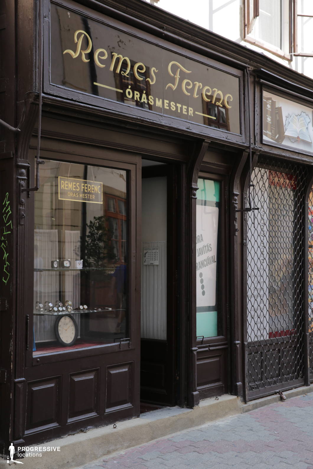 This image captures a charming, old-world watchmaker’s shop with a deep brown wooden facade and gold-lettered signage. The storefront features a glass display showcasing an assortment of classic timepieces, framed by elegant carved details that reflect traditional craftsmanship. The partially open door and adjacent window, protected by a metal security grate, add to the nostalgic, European street scene aesthetic. The worn stone threshold and surrounding aged architecture suggest a long-standing establishment with historical significance. This location is ideal for filming period dramas, mystery films, or character-driven urban stories, offering an intimate and timeless backdrop.