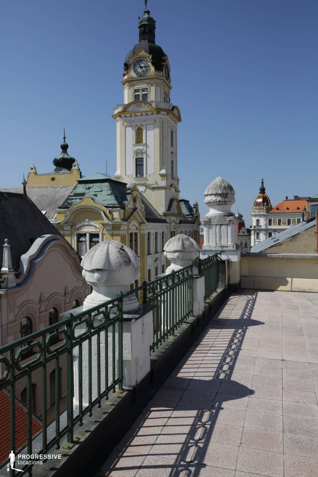 The image showcases a refined rooftop terrace overlooking a cityscape rich in architectural heritage. The ornate metal railing and decorative stone balusters create a sophisticated foreground, leading the eye toward the towering clock tower and surrounding historic buildings adorned with intricate facades, domes, and spires. The soft pastel hues and green patinated rooftops add to the old-world European charm. This location is ideal for filming elegant period dramas, rooftop conversations, or clandestine meetings, offering a stunning panoramic backdrop suited for romance, intrigue, or historical storytelling.