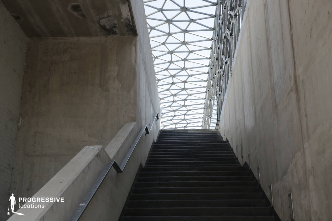 This modern stairway, enclosed by raw concrete walls, leads upward toward a striking geometric ceiling composed of interconnected steel and glass panels. The rigid, angular design creates a contrast between the heavy, industrial materials and the airy, translucent roof structure. The minimalistic railings and embedded lighting elements reinforce the stark, functional aesthetic. The scene is ideal for filming futuristic, dystopian, or psychological thrillers, offering a setting that conveys isolation, transition, or an architectural metaphor for ascension and discovery.