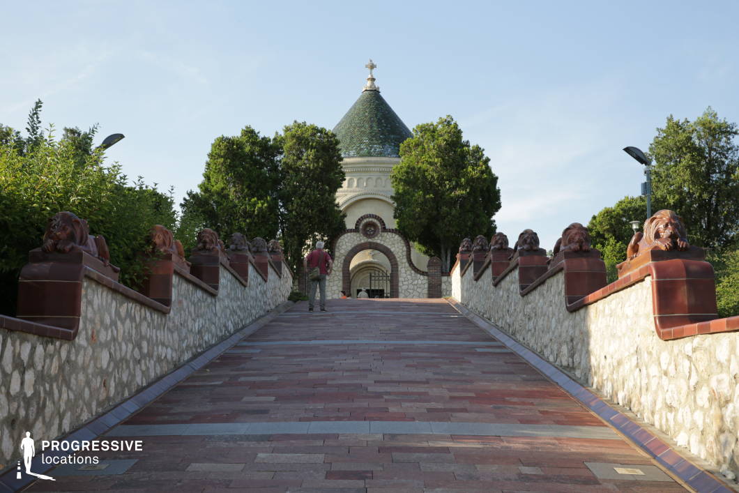 This regal approach to a mausoleum features a stately arched entrance framed by lush greenery and an elegant domed structure in the background. The ascending pathway, flanked by intricately carved lion sculptures on a stone balustrade, adds a sense of grandeur and reverence. The blend of natural stone, red brick, and ceramic tile roofing enhances the historic and ceremonial atmosphere. This location is ideal for filming historical dramas, epic journeys, or reflective narratives, offering a striking and symbolic setting for themes of legacy, honor, and remembrance.