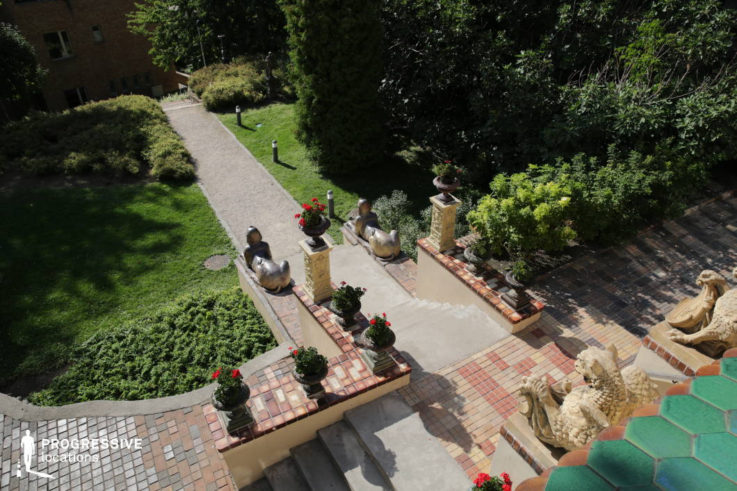 This elegant garden blends natural beauty with decorative artistry, featuring a grand staircase adorned with intricate tilework, sculpted mythical creatures, and ornamental planters filled with vibrant red flowers. The carefully landscaped green space is lined with pathways and trimmed hedges, creating a structured yet inviting atmosphere. The unique bronze and ceramic statues add an artistic, almost whimsical touch, evoking a sense of historical grandeur. This location is ideal for filming period dramas, fantasy films, or cultural documentaries, providing a visually rich backdrop for scenes of artistic inspiration, secret meetings, or poetic reflections in a refined garden setting.