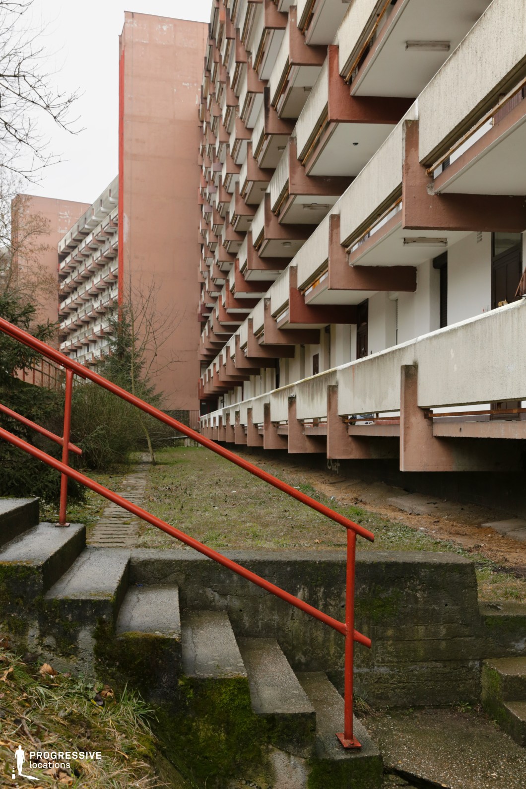 The image captures a striking example of brutalist residential architecture, characterized by a rhythmically repeating series of protruding balconies along a high-rise building clad in muted reddish and off-white tones. The rigid linearity of the concrete elements, combined with the sharp shadows cast by each overhanging form, creates a visually dynamic facade. A red metal handrail leads up a moss-covered concrete stairway in the foreground, introducing a vivid geometric contrast that directs the viewer’s eye toward the imposing structure. The overall atmosphere is one of urban density and functional design, with a focus on raw textures and modular repetition. This location is ideal for filming urban dramas, dystopian narratives, or architectural documentaries, and works especially well for scenes that require a dense residential backdrop with a strong visual identity.