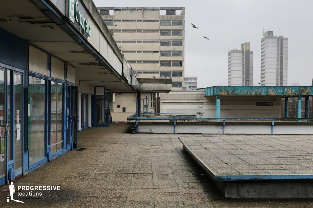 This location showcases a Brutalist-style urban terrace characterized by raw concrete surfaces, sharp geometric lines, and functional design elements. The foreground features a large open paved area with raised platforms and low blue railings, while the building on the left displays a repetitive pattern of glass shopfronts framed in blue metal. The architecture includes weathered concrete canopies and angular overhangs, creating a sense of depth and layering. In the background, multiple high-rise buildings with uniform window arrangements reinforce the location’s mid-to-late 20th century architectural aesthetic. The muted grey and blue palette enhances its stark, utilitarian atmosphere. This space is ideal for filming gritty urban scenes, dystopian or retro-futuristic narratives, or as a backdrop for music videos and fashion shoots requiring a raw, industrial tone.